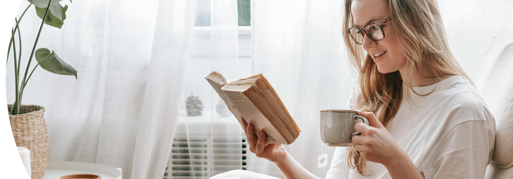 Junge Frau sitzt mit Lesebrille im Sessel, hält eine Tasse und liest ein Buch