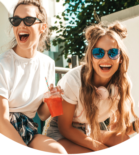 Zwei Frauen, die lachend zusammen auf einer Bank sitzen. Beide trangen T-Shirts und Sonnenbrillen