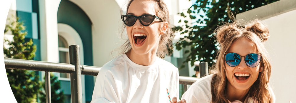 Zwei Frauen, die lachend zusammen auf einer Bank sitzen. Beide trangen T-Shirts und Sonnenbrillen